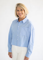 A smiling blonde woman wearing a light blue and white vertically striped button-down shirt with a chest pocket, paired with white pants, against a plain white studio background.