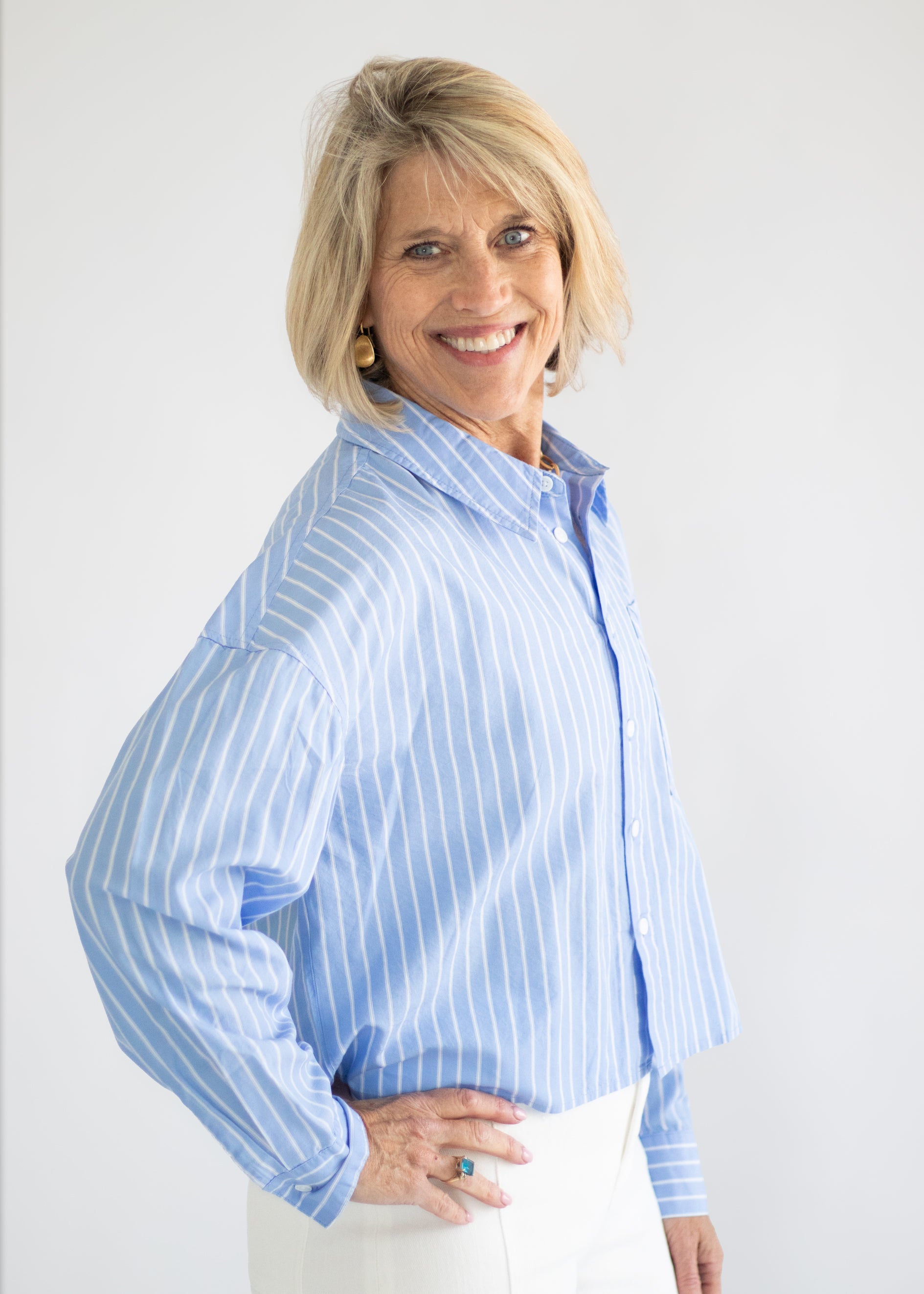 A smiling blonde woman wearing a light blue and white vertically striped button-down shirt and white pants, standing against a plain light gray background.