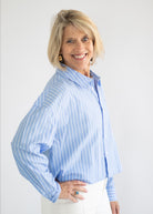 A smiling blonde woman wearing a light blue and white vertically striped button-down shirt and white pants, standing against a plain light gray background.