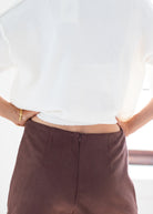 Close-up back view of a person wearing a loose white top and dark brown high-waisted trousers, with hands in pockets and a gold bracelet visible on one wrist.