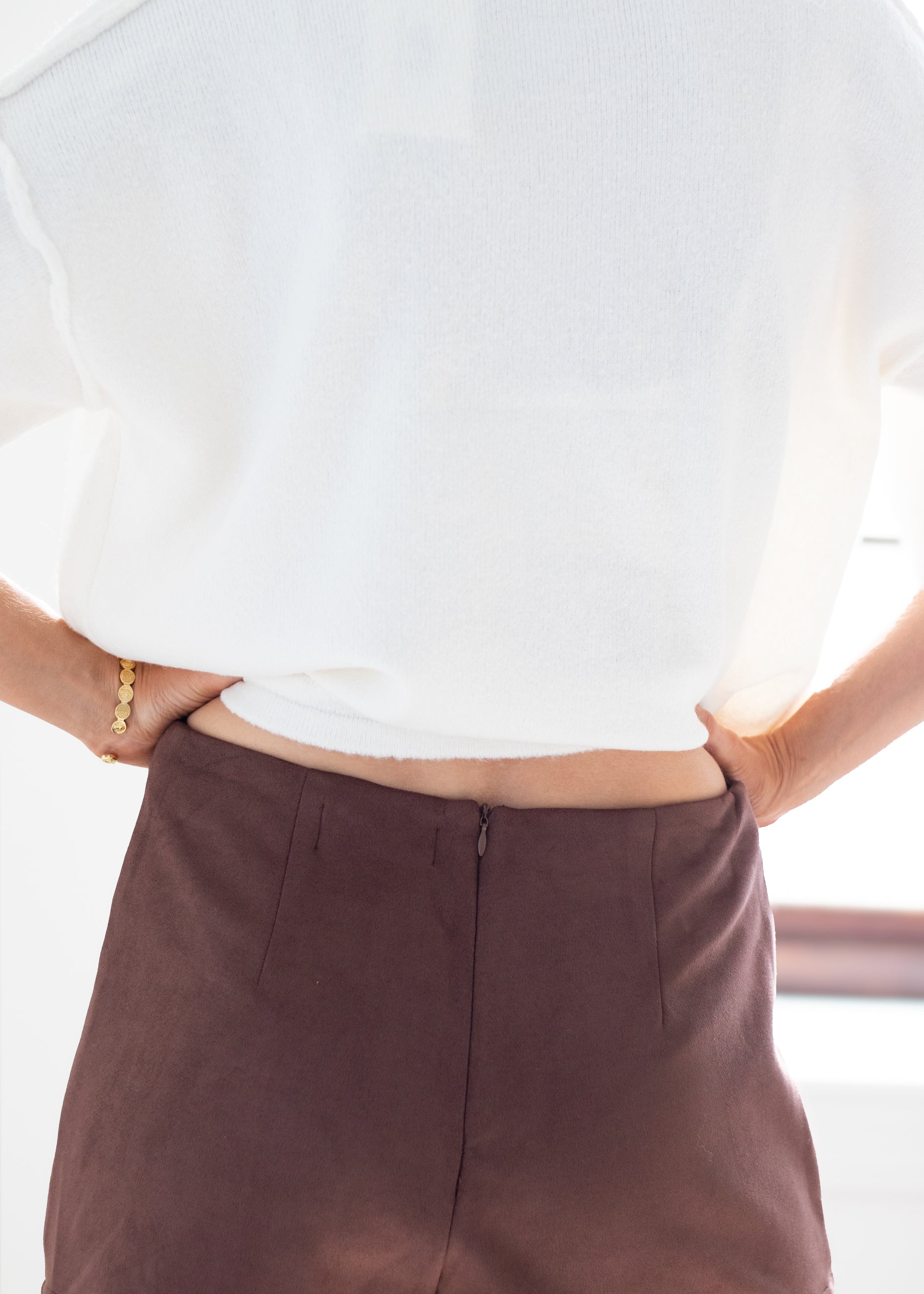 Close-up back view of a person wearing a loose white top and dark brown high-waisted trousers, with hands in pockets and a gold bracelet visible on one wrist.
