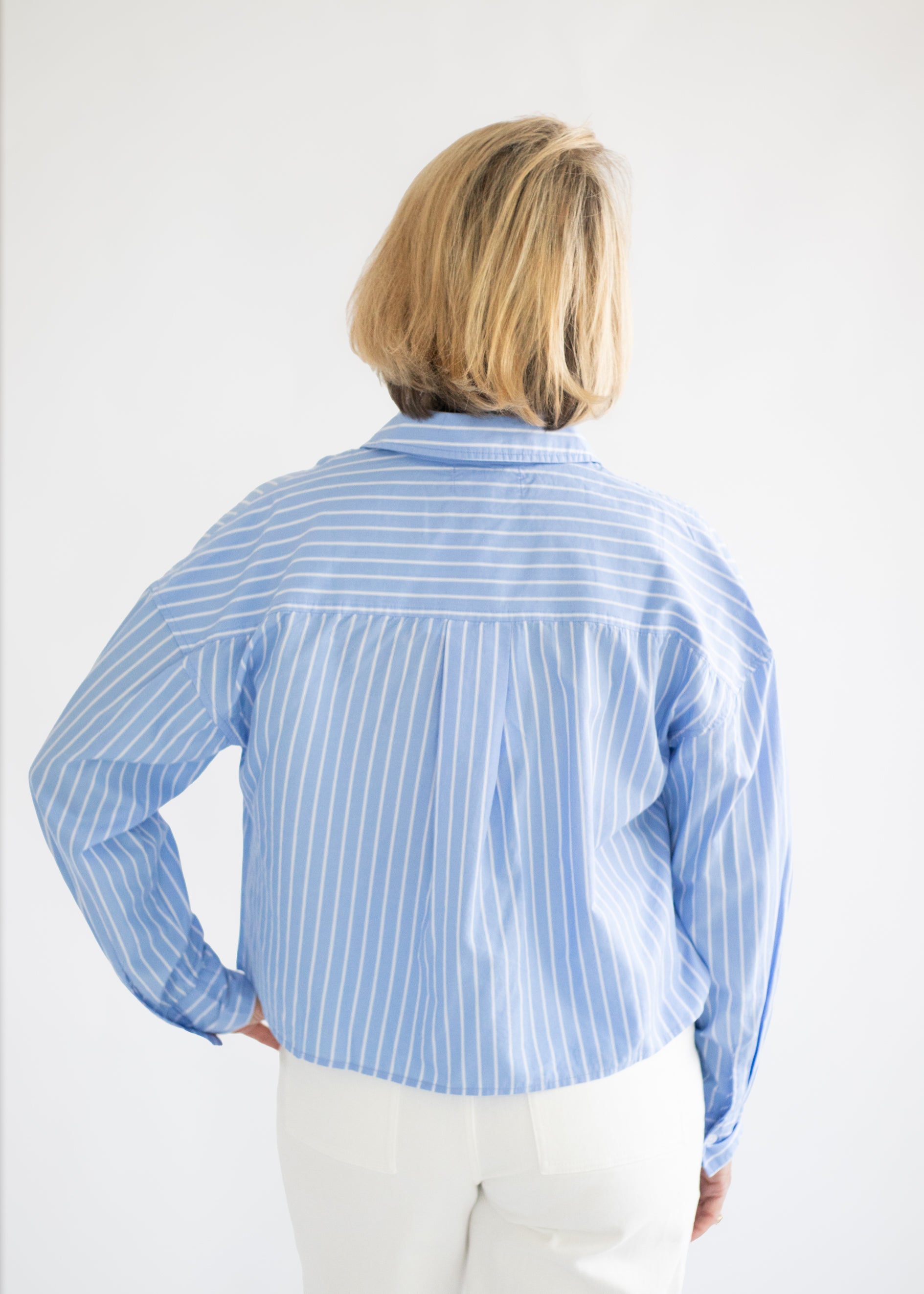 A woman viewed from behind wearing a light blue and white vertically striped button-down shirt and white pants, set against a plain white background.