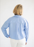 A woman viewed from behind wearing a light blue and white vertically striped button-down shirt and white pants, set against a plain white background.