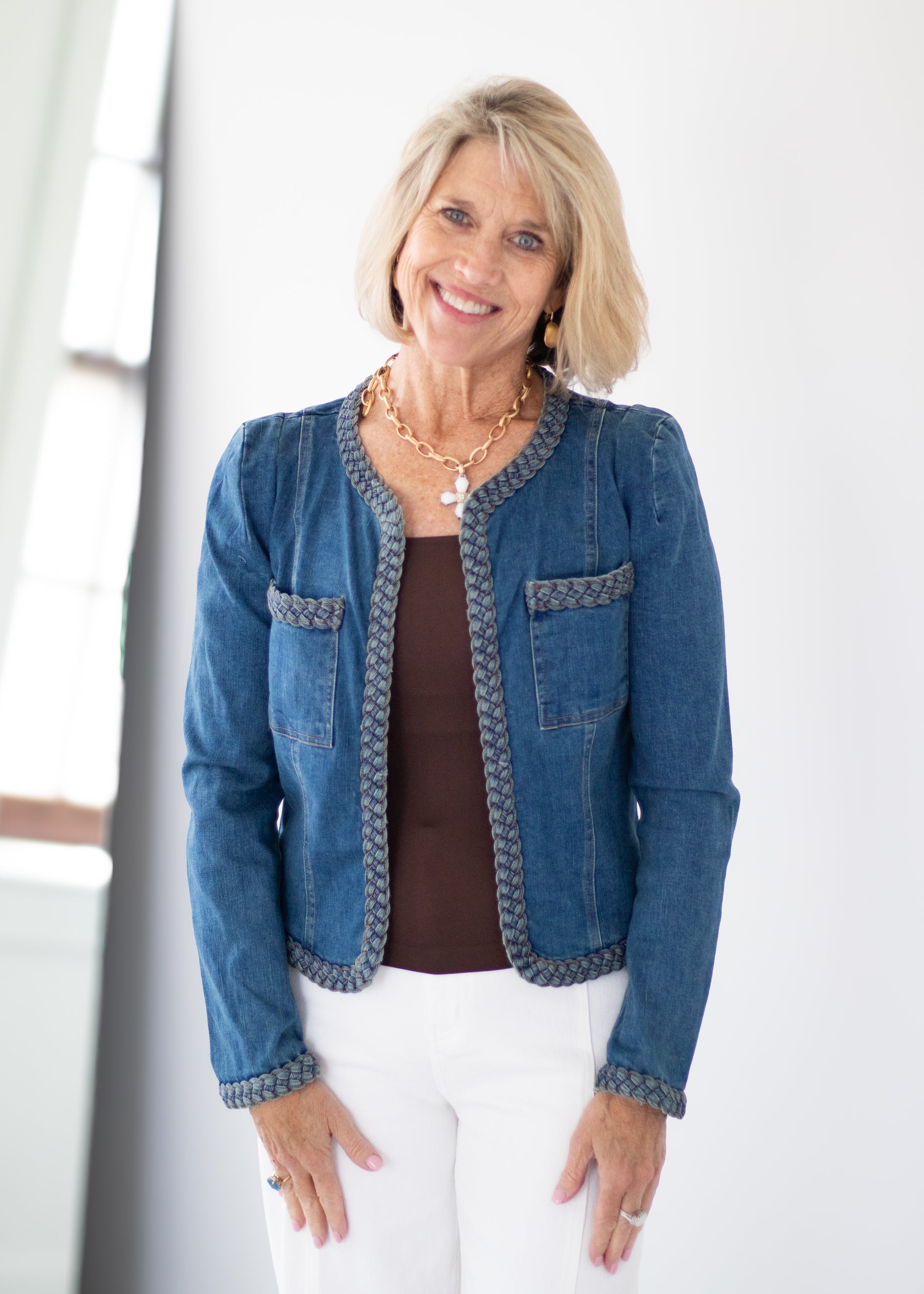 A smiling blonde woman wearing a blue denim jacket with patterned trim over a brown top and white pants, standing in a bright indoor setting.