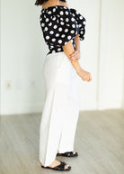 Person wearing a black and white polka dot top with white pants in a minimal indoor setting.