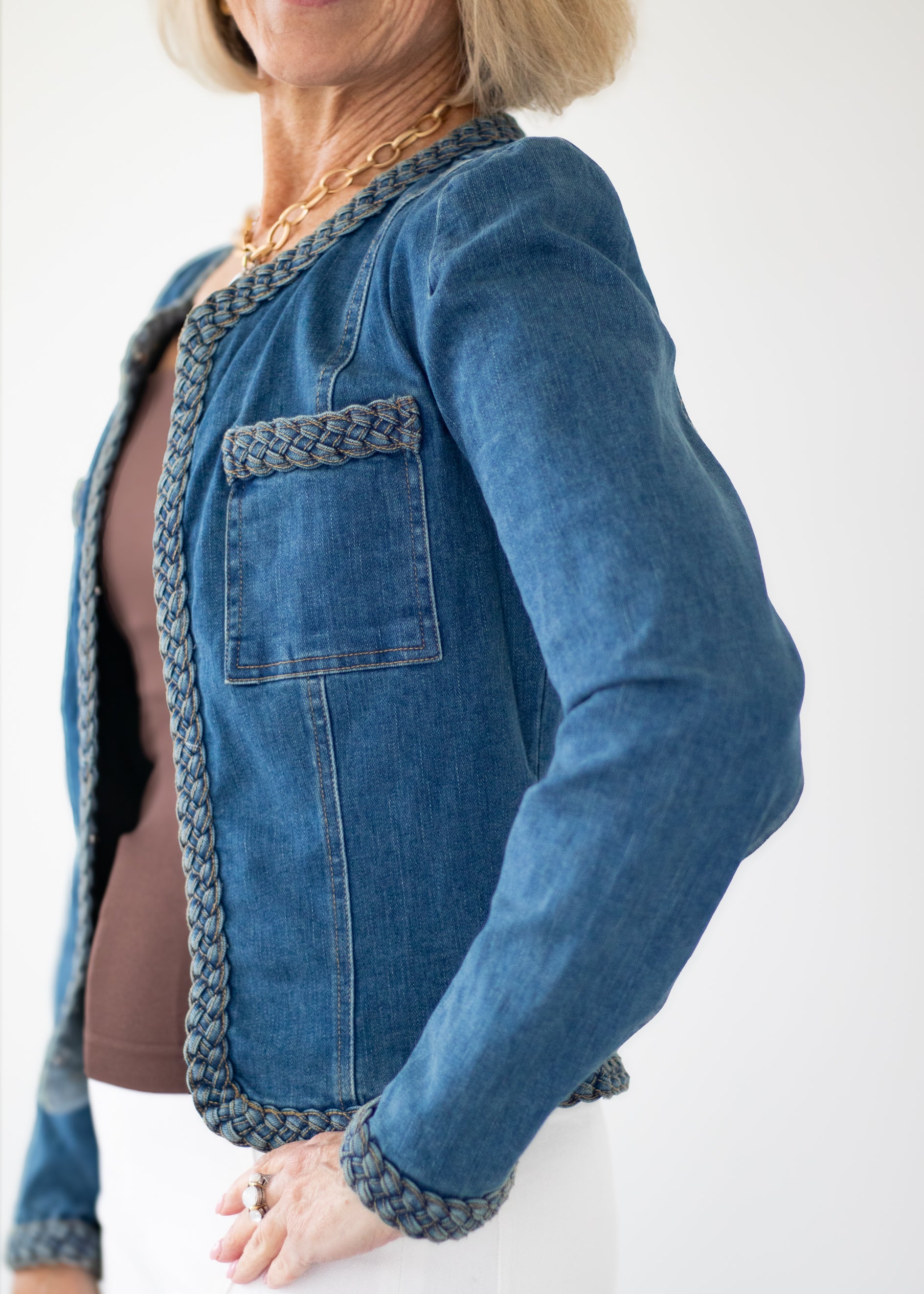 A woman wearing a blue denim jacket with braided trim, styled over a brown top and white pants against a plain white background.