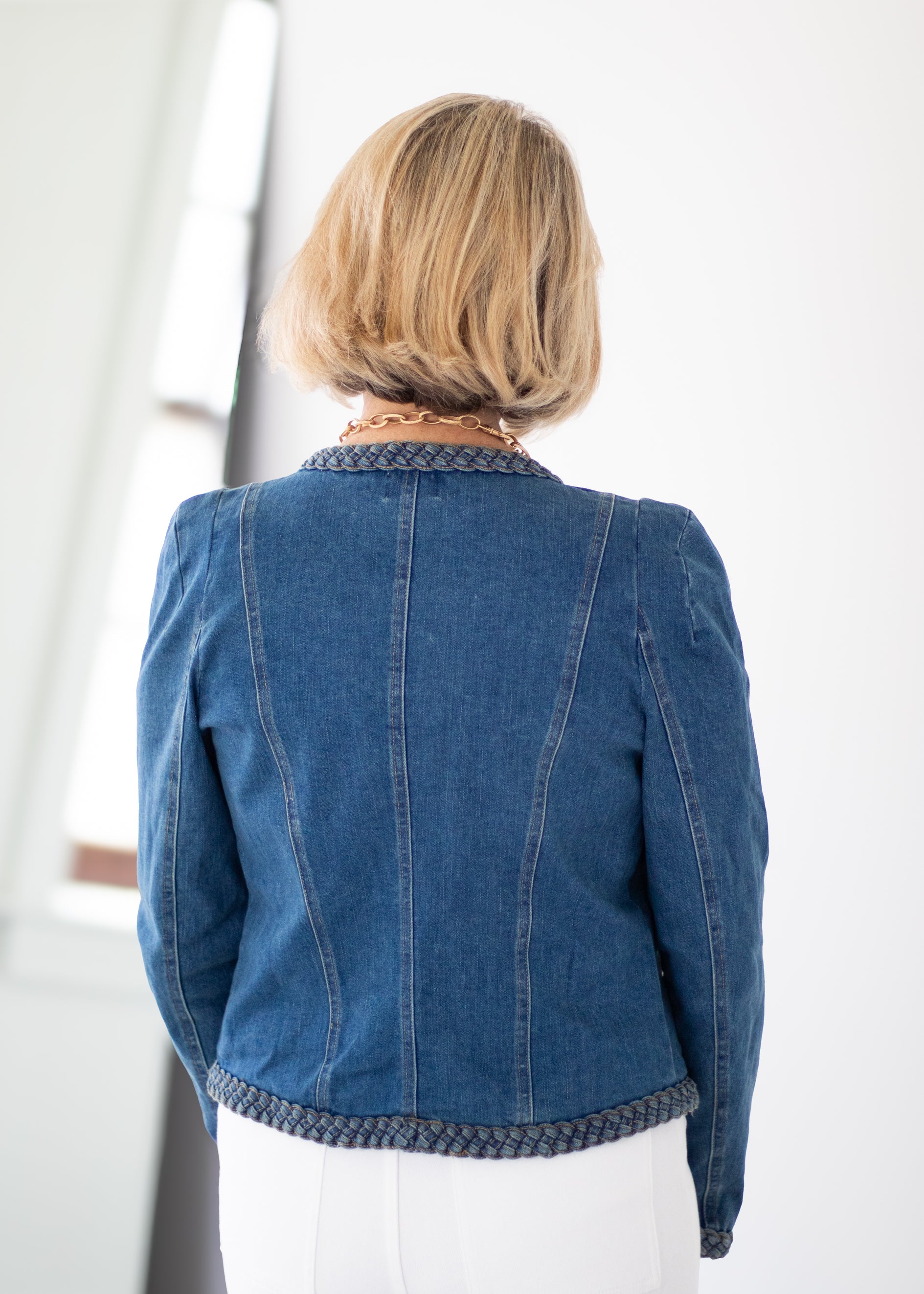 A woman viewed from behind wearing a blue denim jacket with patterned trim on the collar and hem, paired with white pants in a bright indoor setting.