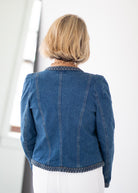 A woman viewed from behind wearing a blue denim jacket with patterned trim on the collar and hem, paired with white pants in a bright indoor setting.