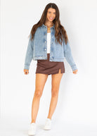 A woman modeling a light-wash blue denim jacket over a white top and brown mini skirt, paired with white sneakers, against a plain white studio background.