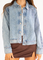 A woman wearing a light-wash blue denim jacket over a white top and brown mini skirt, shown from the shoulders to the upper thighs against a plain light background.