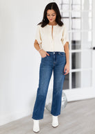 Woman wearing a cream blouse and blue jeans with white boots in a minimalistic setting.