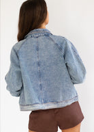 A woman shown from the back wearing a light blue denim jacket and brown shorts against a plain white background.