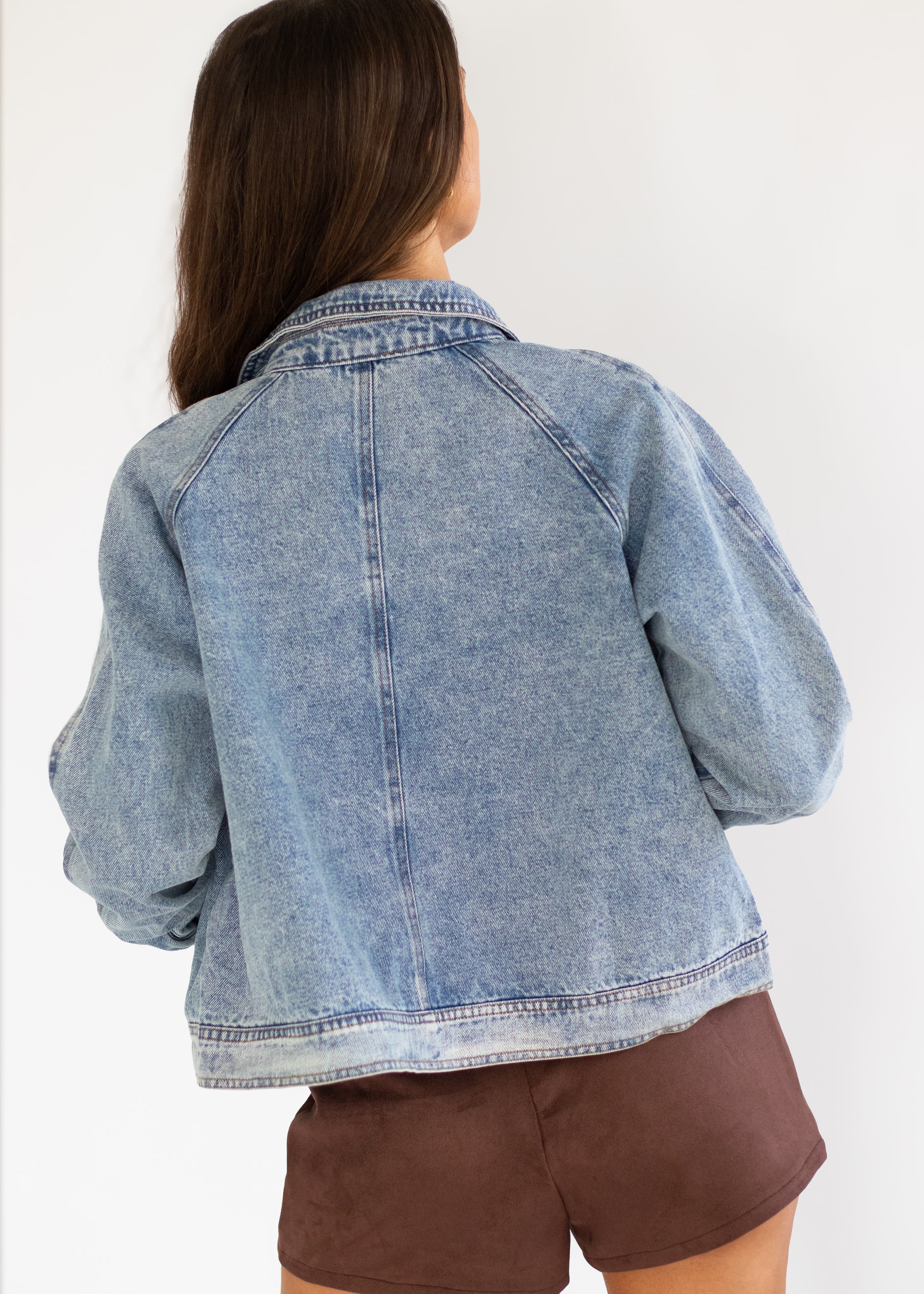A woman shown from the back wearing a light blue denim jacket and brown shorts against a plain white background.