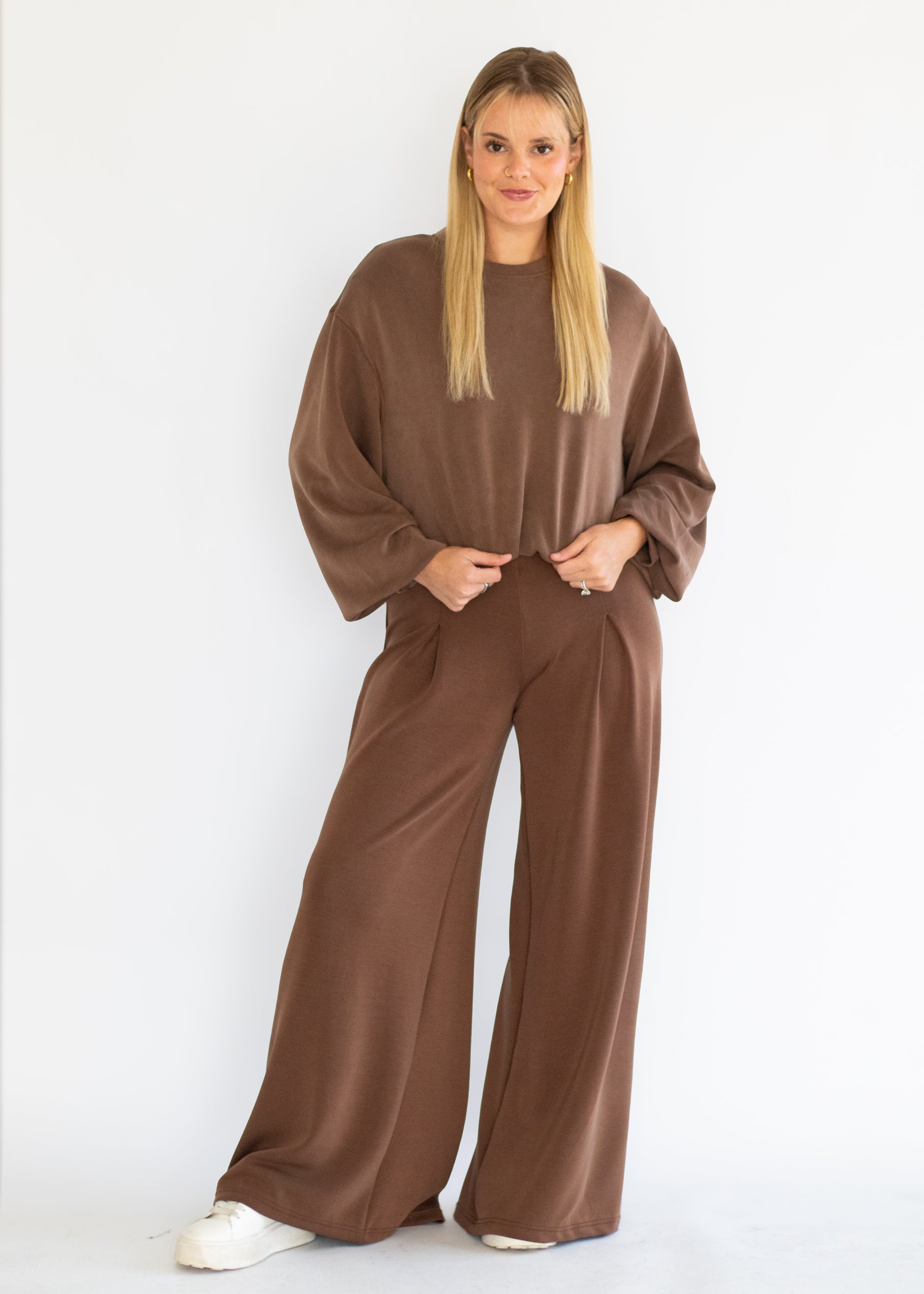 A woman modeling a matching brown lounge set consisting of a long-sleeve top and wide-leg pants, paired with white sneakers against a plain white studio background.
