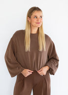 A blonde woman wearing a matching brown lounge set, featuring a loose long-sleeve top and coordinating pants, standing against a plain white background.