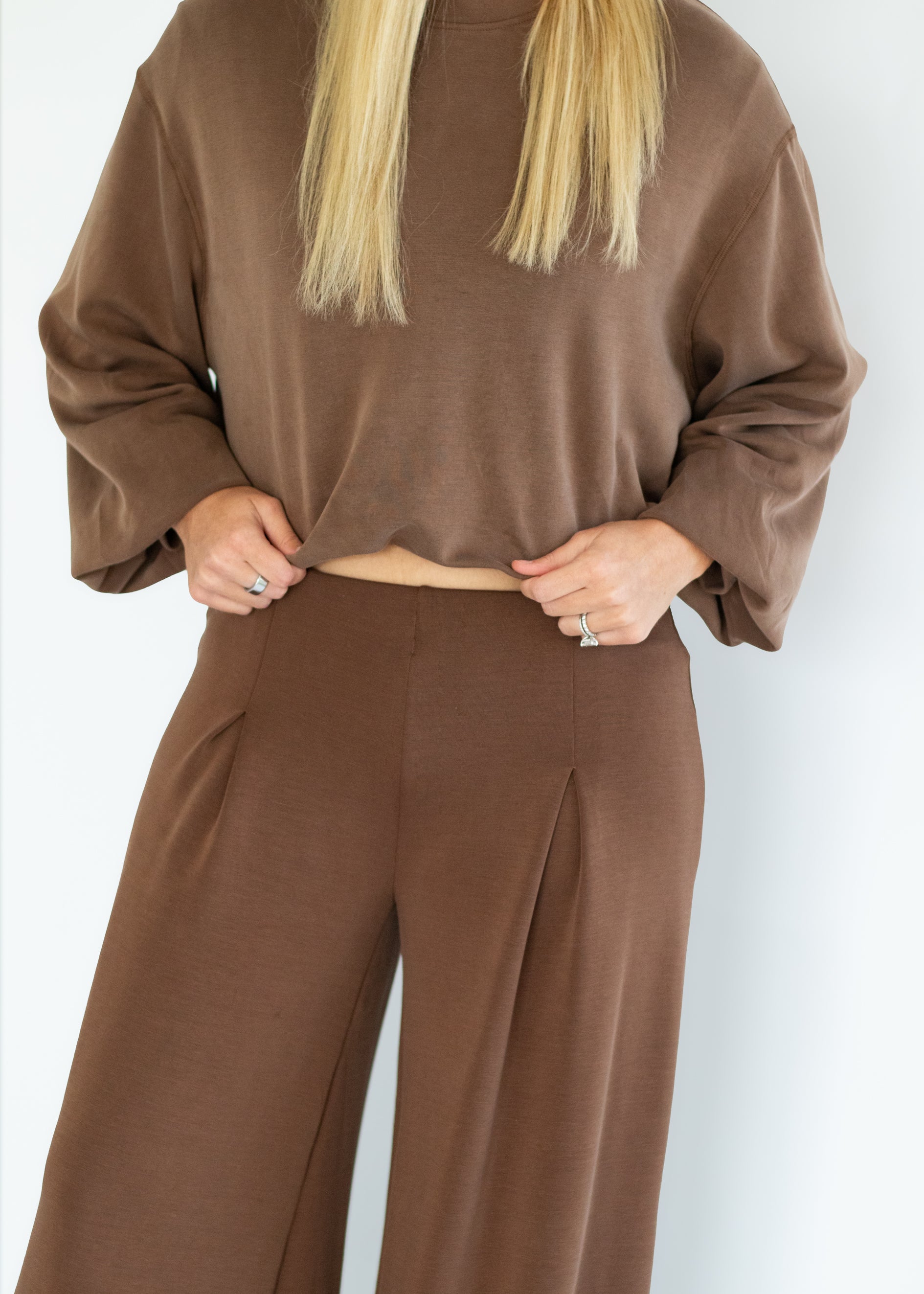 A woman wearing a brown long-sleeve cropped top and matching brown wide-leg trousers, shown from the shoulders down against a plain light background.