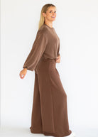 A woman modeling a matching two-piece lounge set in a solid brown color, featuring a long-sleeve top and wide-leg pants, photographed against a plain white studio background.