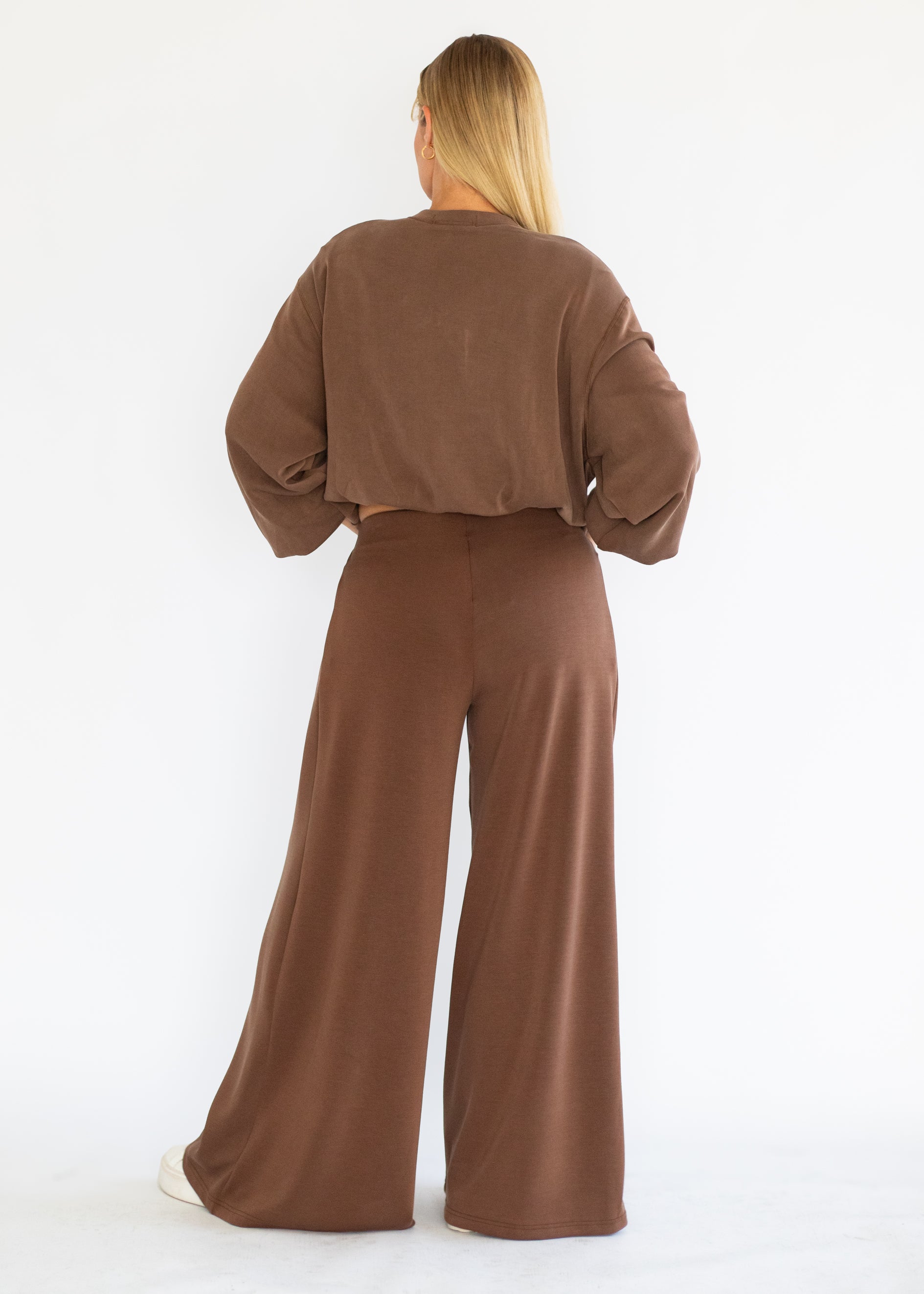 A woman viewed from behind wearing a matching brown long-sleeve cropped top and wide-leg pants set, standing against a plain white studio background.