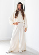 A woman modeling a matching cream two-piece lounge set consisting of a cropped long-sleeve top and high-waisted wide-leg pants, paired with white sneakers in a bright, minimalist room.