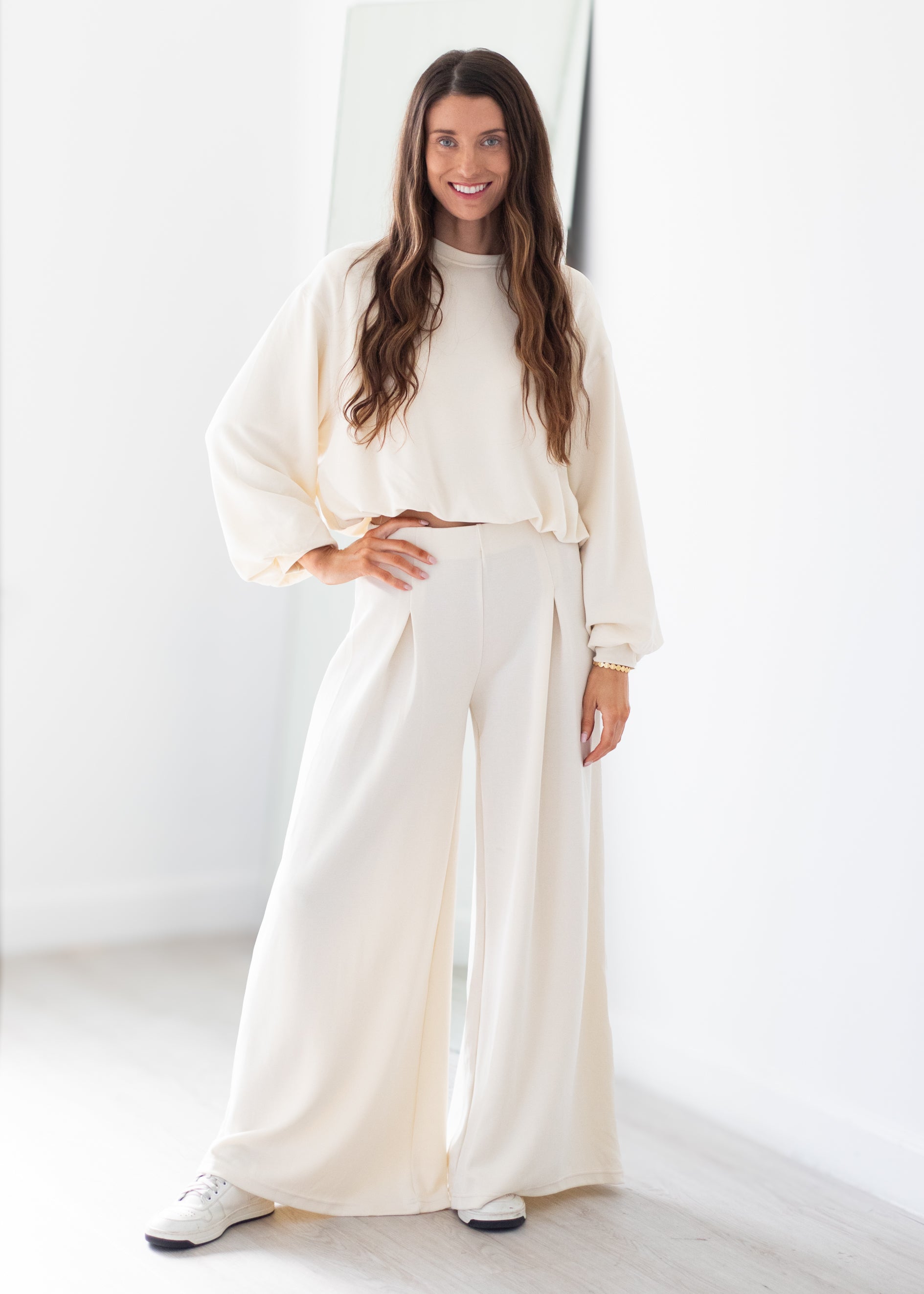 A woman modeling a matching cream two-piece lounge set consisting of a cropped long-sleeve top and high-waisted wide-leg pants, paired with white sneakers in a bright, minimalist room.