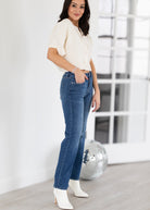 Woman wearing a white top and blue jeans with white shoes in a minimalistic room.