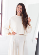 A woman with long brown hair smiling and posing in a bright, minimalist room. She is wearing a cream-colored cropped sweatshirt with balloon sleeves and matching cream wide-leg trousers.