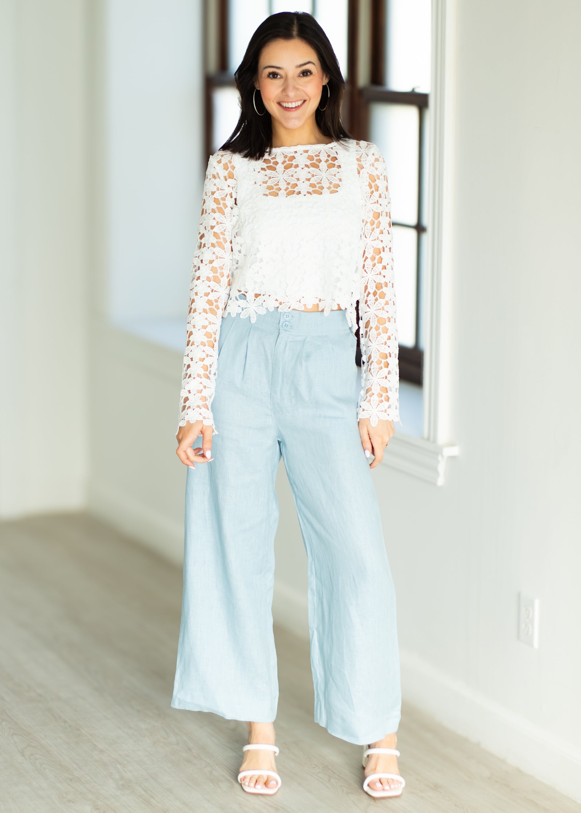 Woman wearing a white lace top and light blue pants in a bright room.