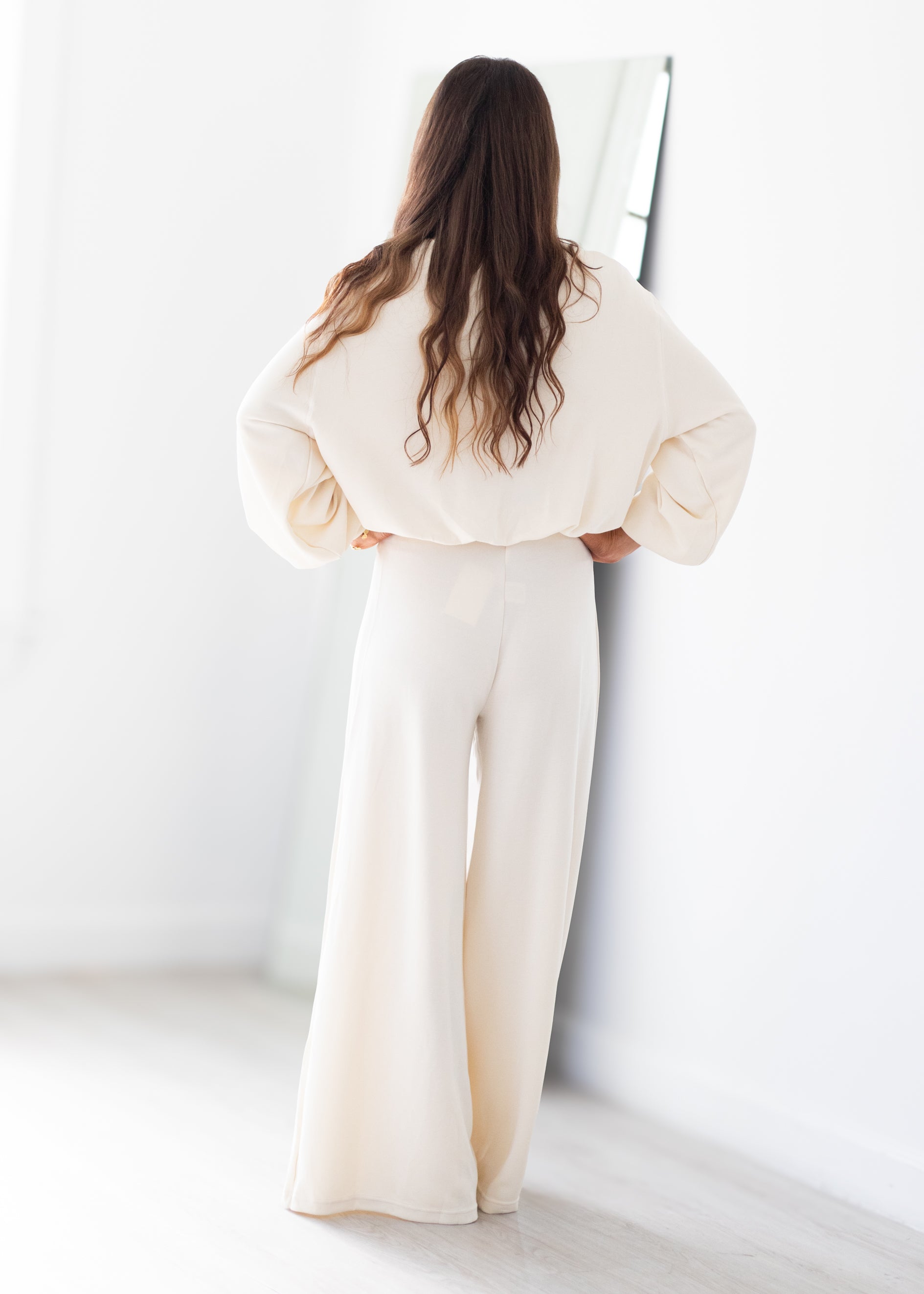 A woman with long brown hair is shown from behind wearing a cream-colored two-piece lounge set consisting of a cropped long-sleeve top and matching wide-leg pants, standing in a bright, minimalist room.