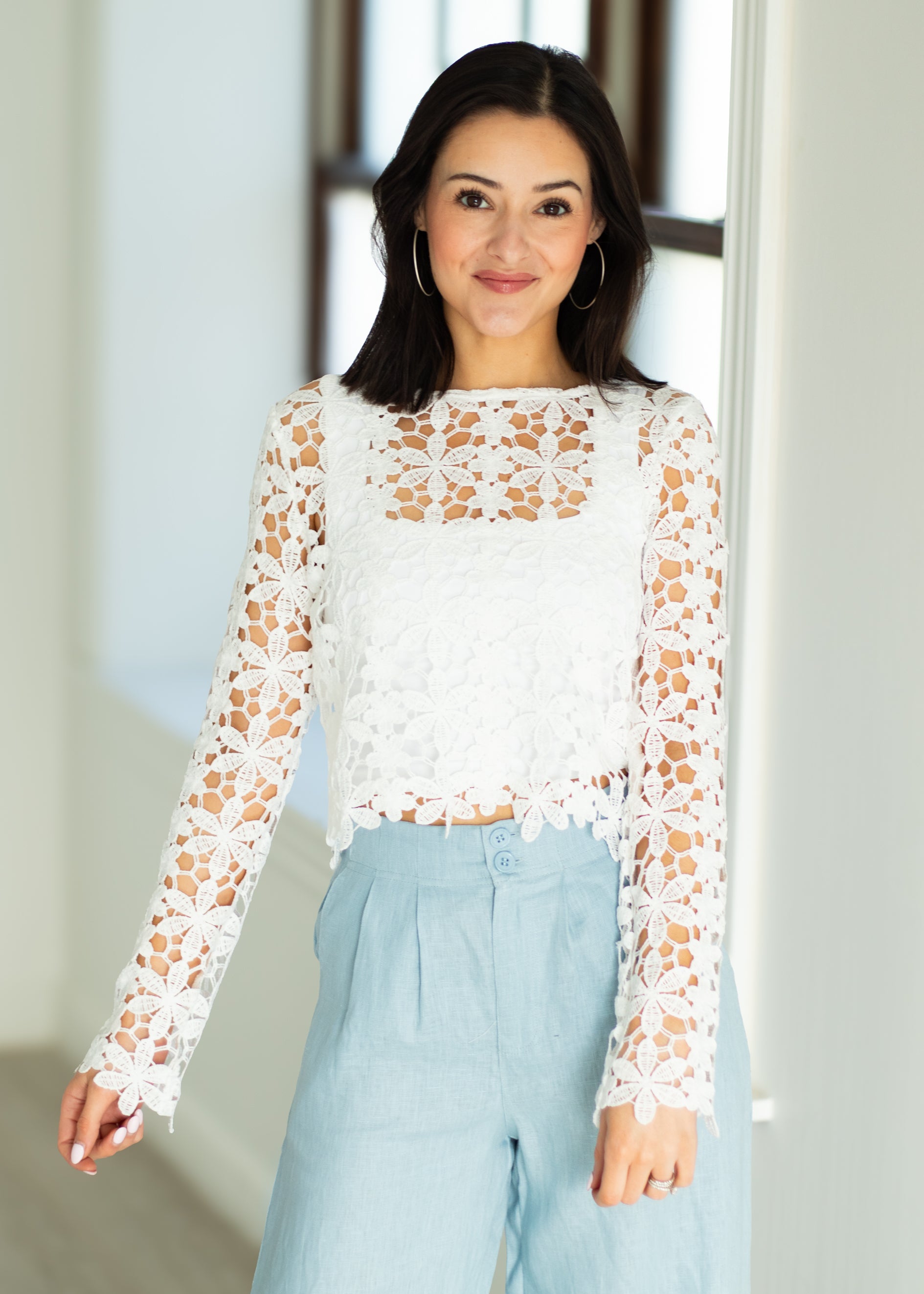 Woman wearing a white lace top and light blue pants indoors.
