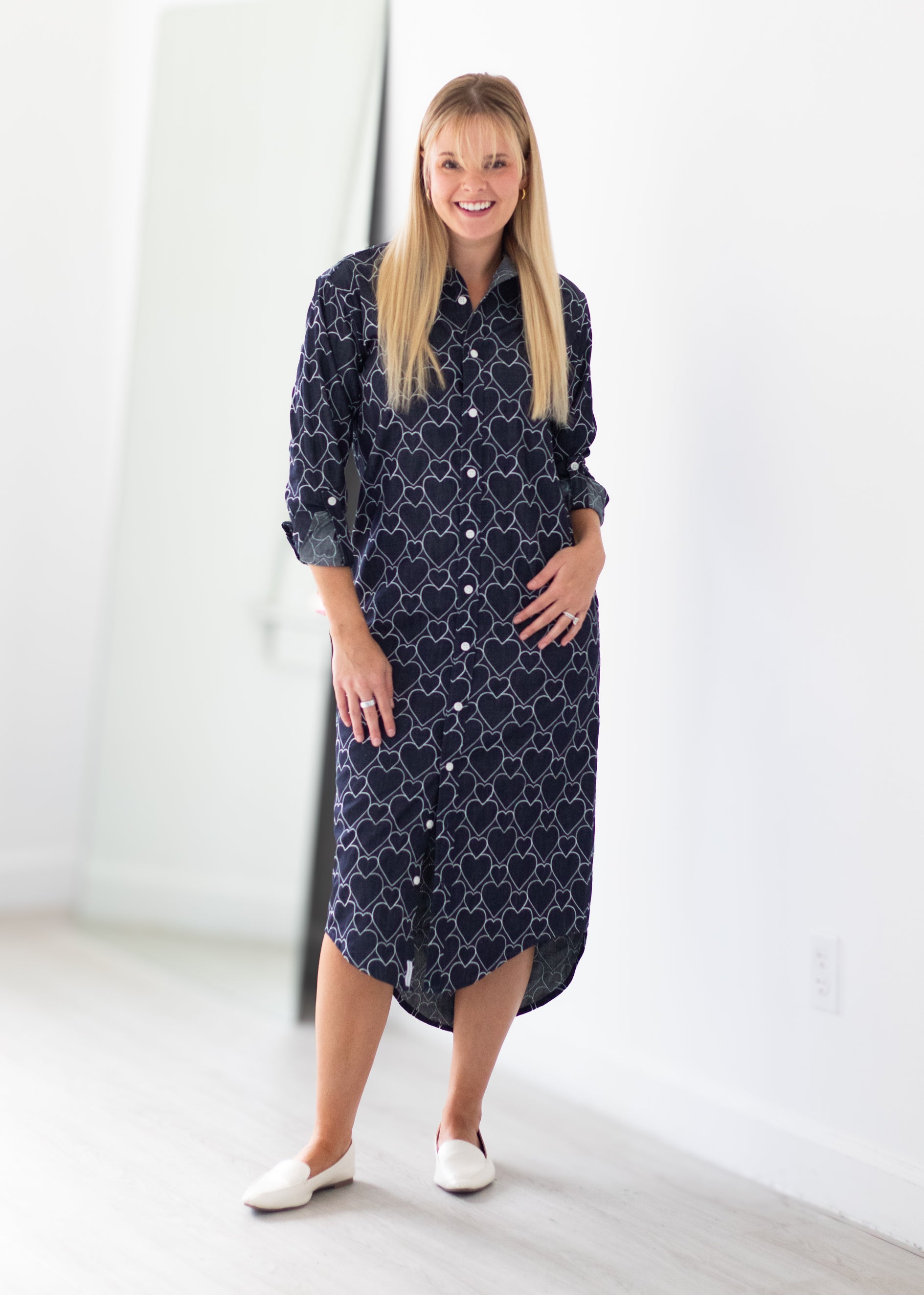 A woman stands in a bright, minimalist room wearing a navy blue button-up shirt dress with a white geometric pattern, paired with white flat shoes.