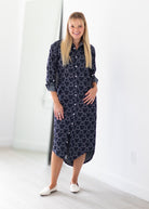 A woman stands in a bright, minimalist room wearing a navy blue button-up shirt dress with a white geometric pattern, paired with white flat shoes.