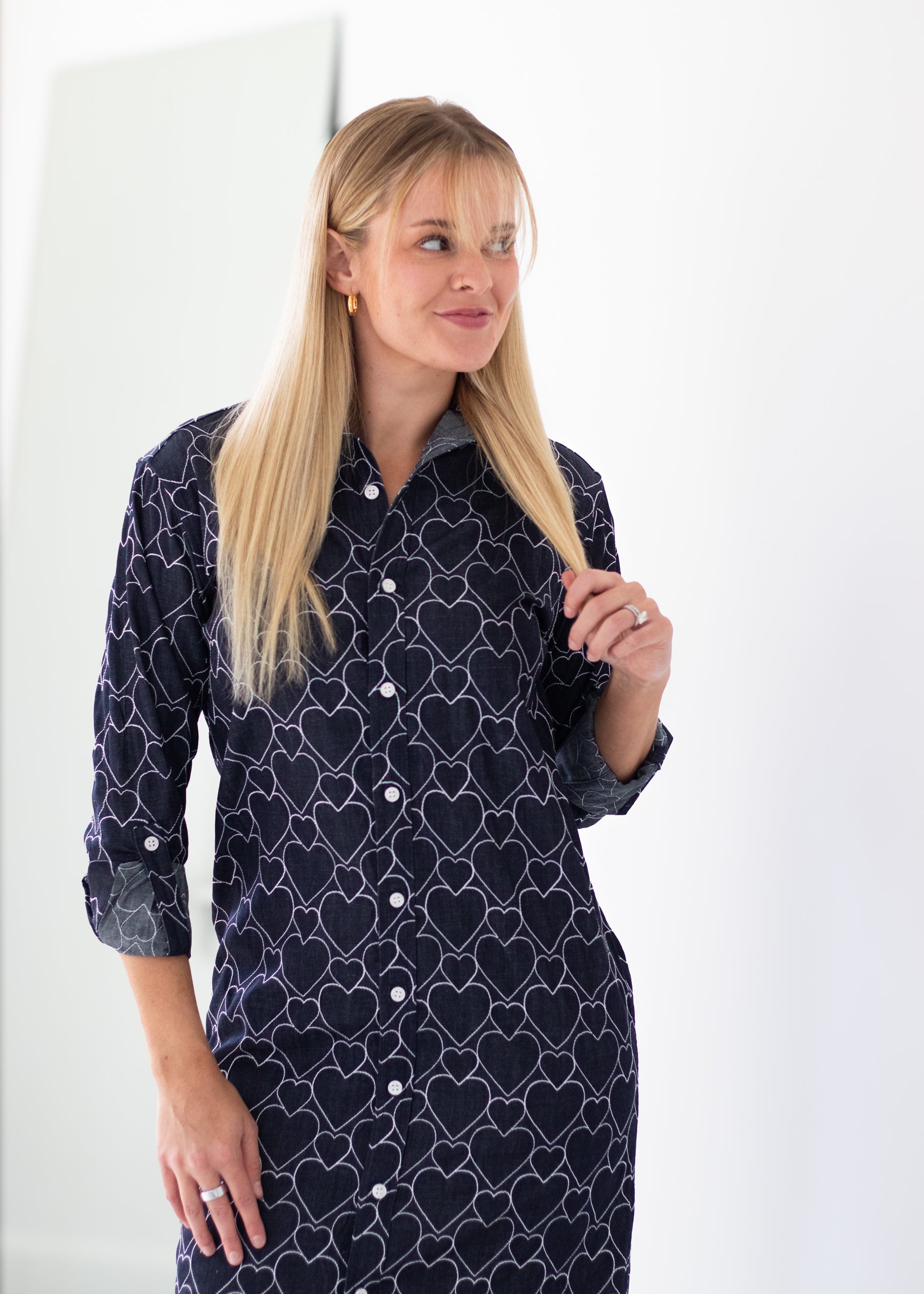 A blonde woman wearing a dark navy long-sleeve button-up shirt dress with a white geometric heart pattern, standing against a bright white background.
