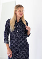 A blonde woman wearing a dark navy long-sleeve button-up shirt dress with a white geometric heart pattern, standing against a bright white background.