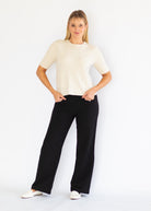 A woman modeling a cream short-sleeve knit top and black wide-leg trousers, standing against a plain white studio background.