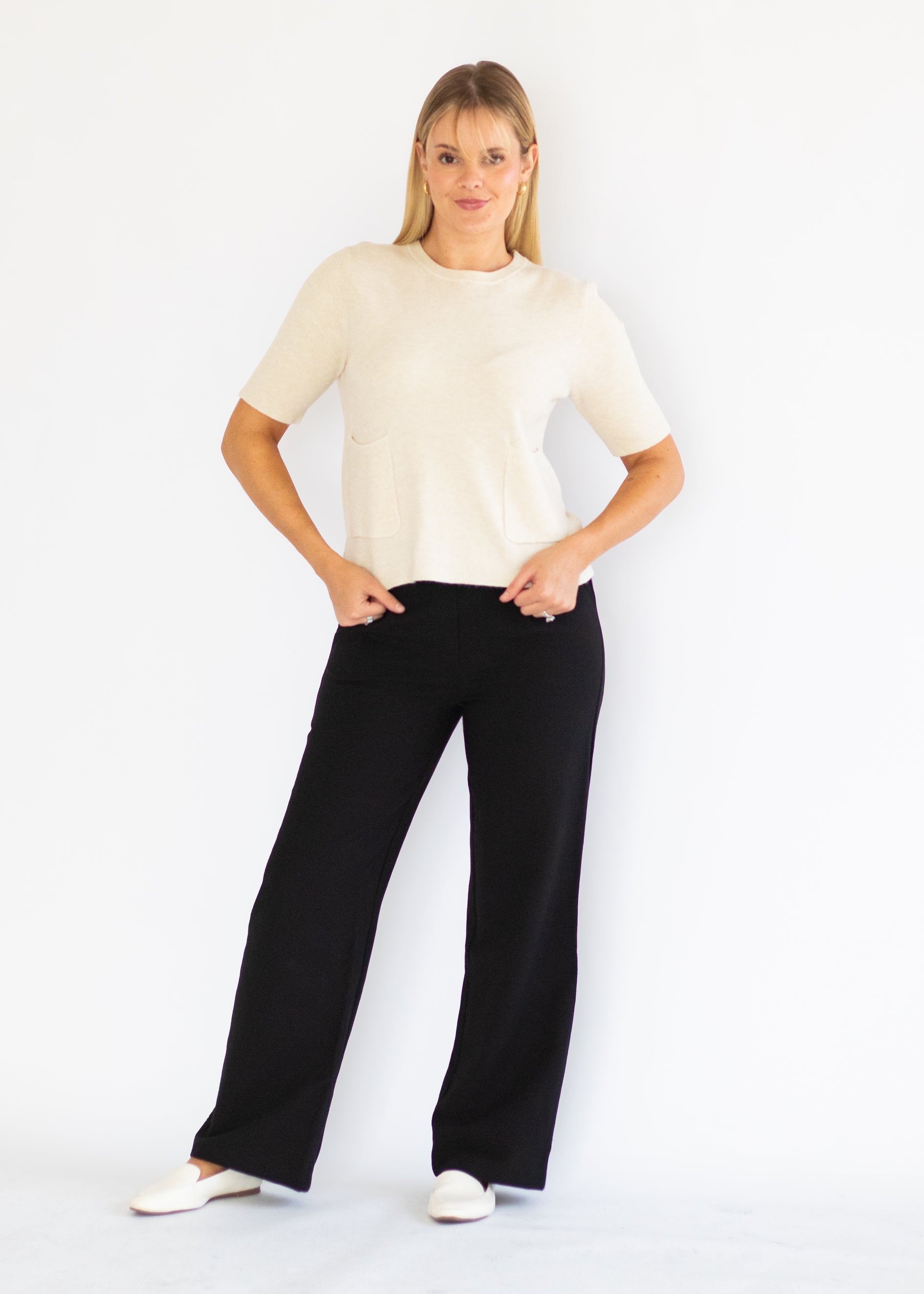 A woman modeling a cream short-sleeve knit top and black wide-leg trousers, standing against a plain white studio background.