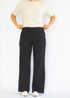 A model wearing black wide-leg trousers with a cream short-sleeve knit top and white slip-on shoes, standing against a plain white studio background.