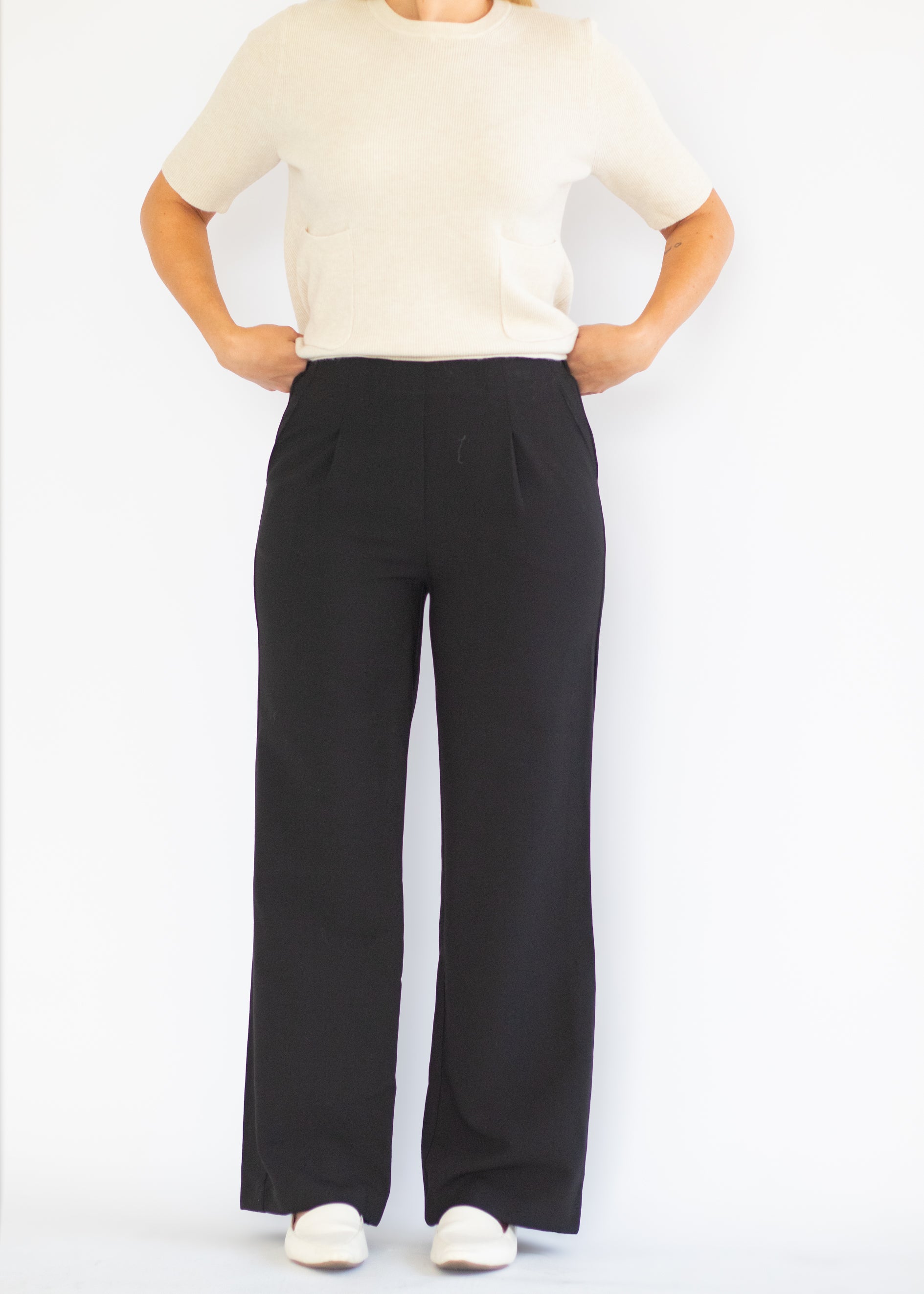 A model wearing black wide-leg trousers with a cream short-sleeve knit top and white slip-on shoes, standing against a plain white studio background.