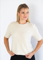 A woman modeling a short-sleeve knit top in a warm off-white or cream color, paired with black pants, against a plain white studio background.