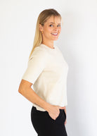 A woman with blonde hair is shown from the side, wearing a short-sleeve cream knit top and black pants against a plain white background.