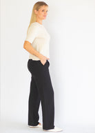 A woman standing in profile wearing a cream short-sleeve knit top and black wide-leg trousers, photographed against a plain white studio background.