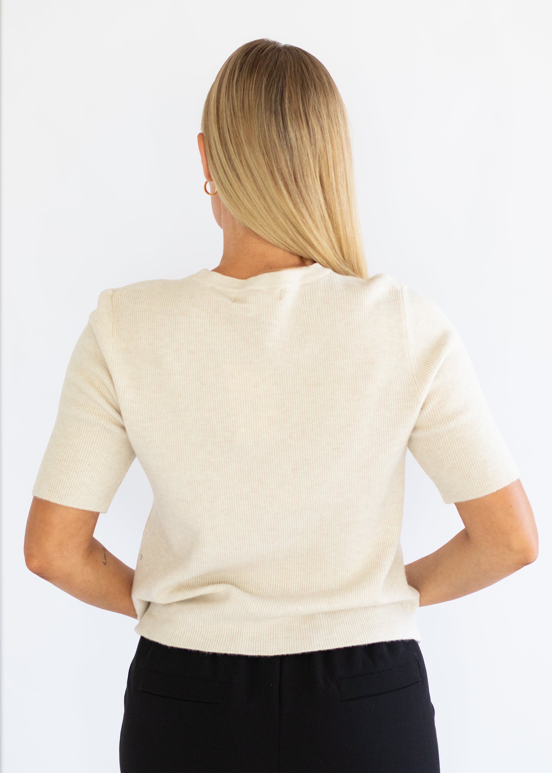 A woman viewed from the back wearing a short-sleeve cream knit top and black pants against a plain white background.