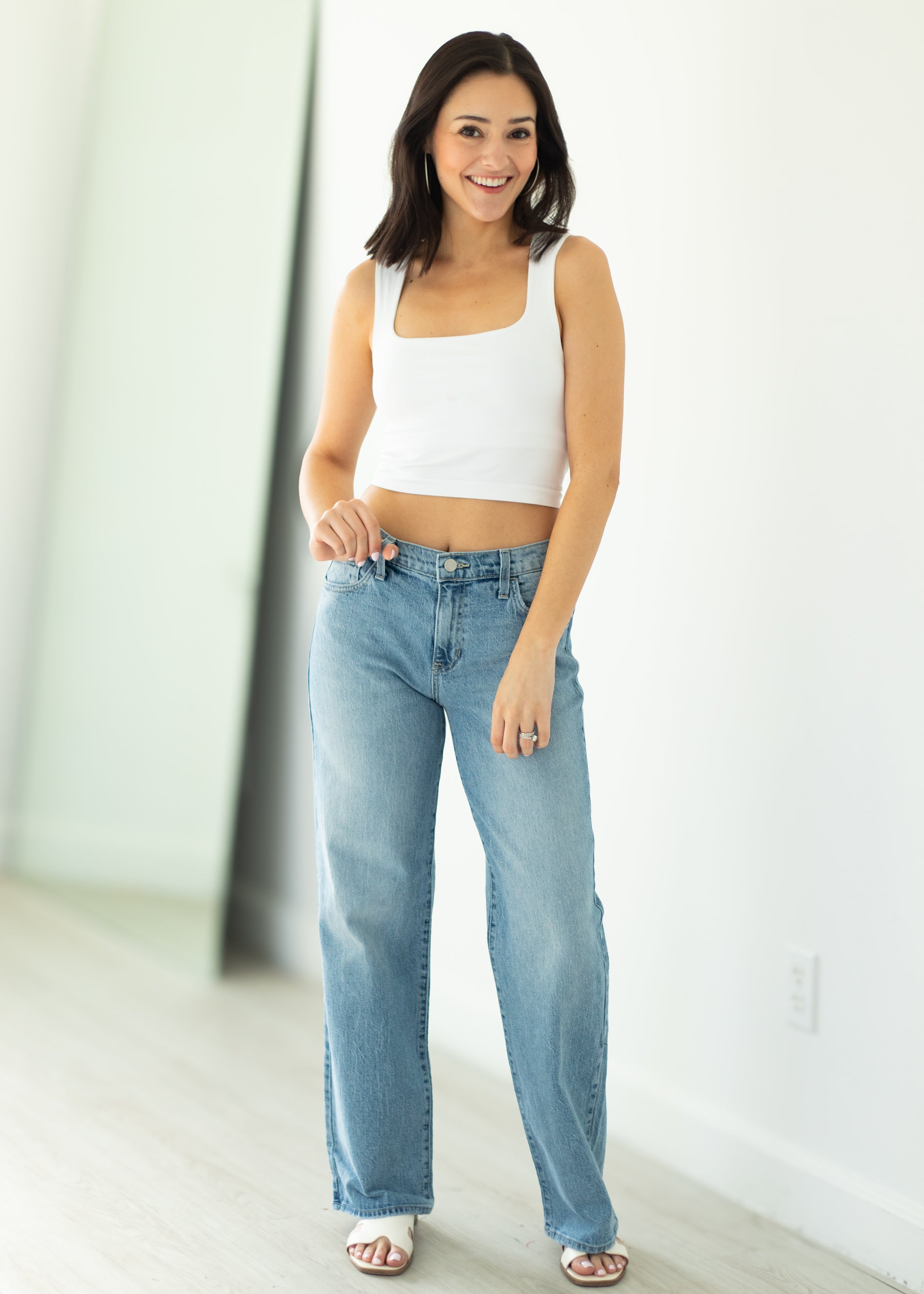Woman wearing a white tank top and light blue jeans standing against a white wall.