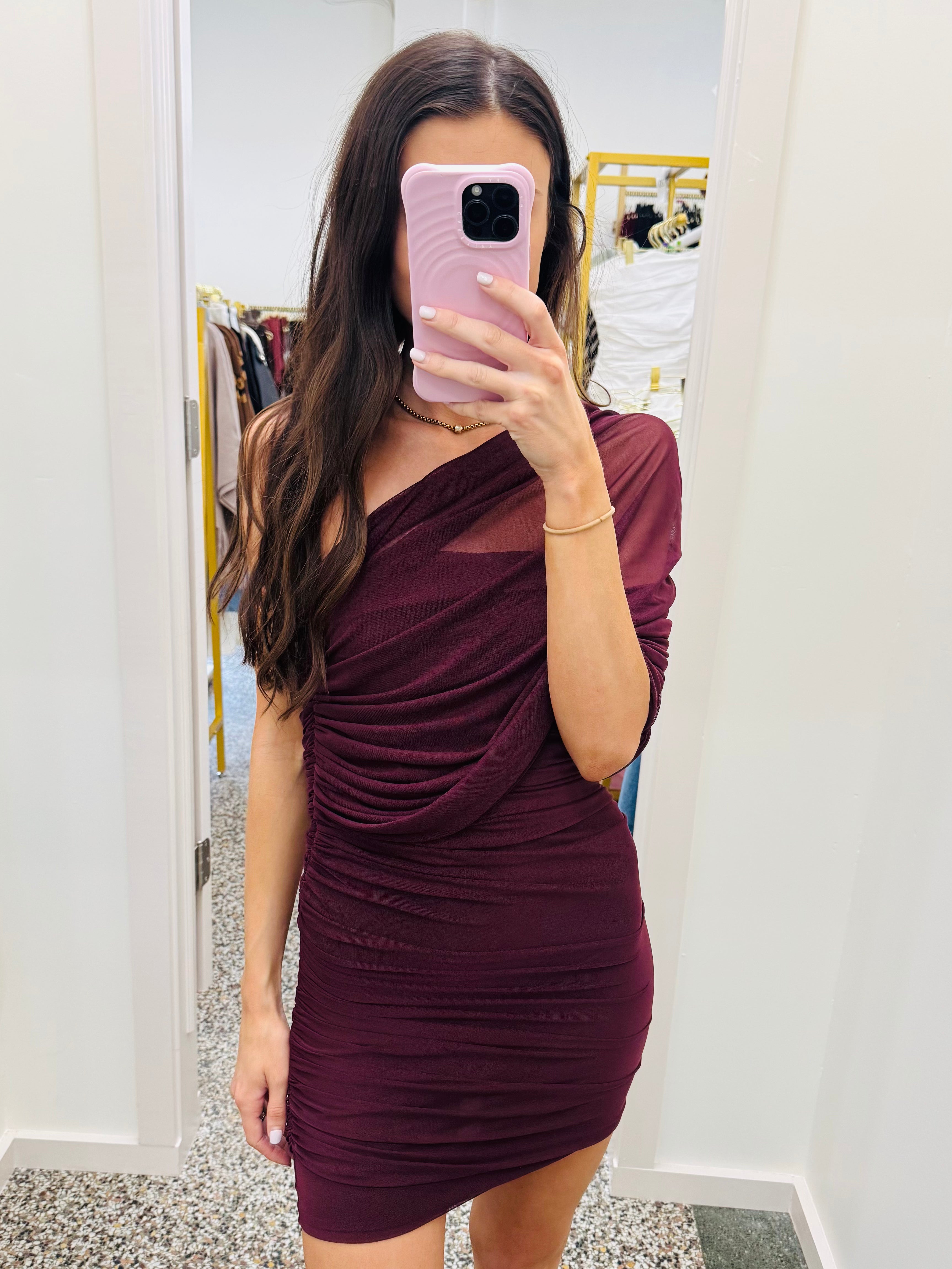 A woman takes a mirror selfie in a fitting room wearing a ruched, one-shoulder mini dress in deep burgundy. She is holding a light pink smartphone.