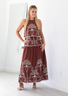 A woman modeling a sleeveless rust-brown maxi dress with intricate white embroidery and lace trim, standing in a bright, minimalist room with white walls and light wood floors.