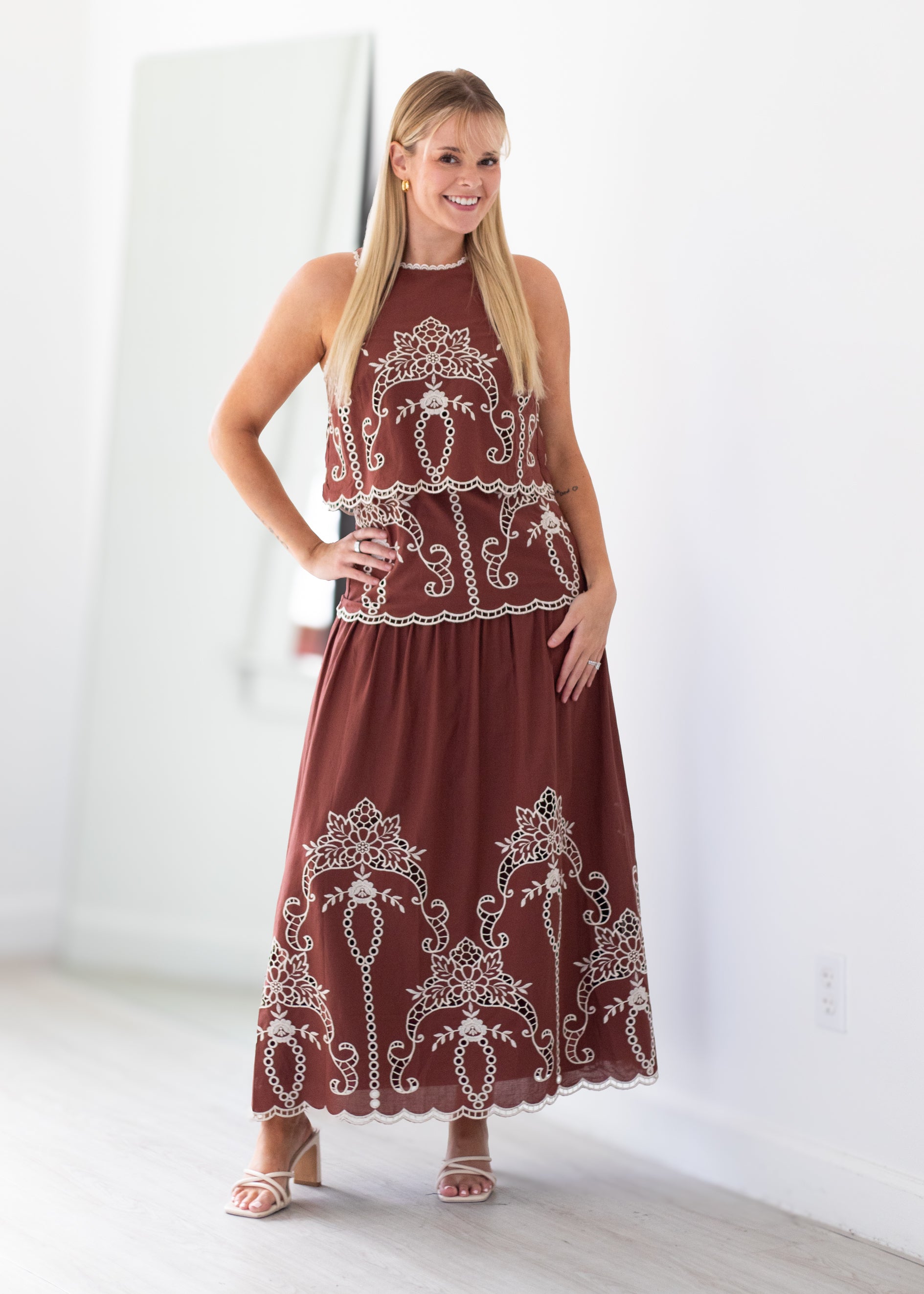 A woman modeling a sleeveless rust-brown maxi dress with intricate white embroidery and lace trim, standing in a bright, minimalist room with white walls and light wood floors.