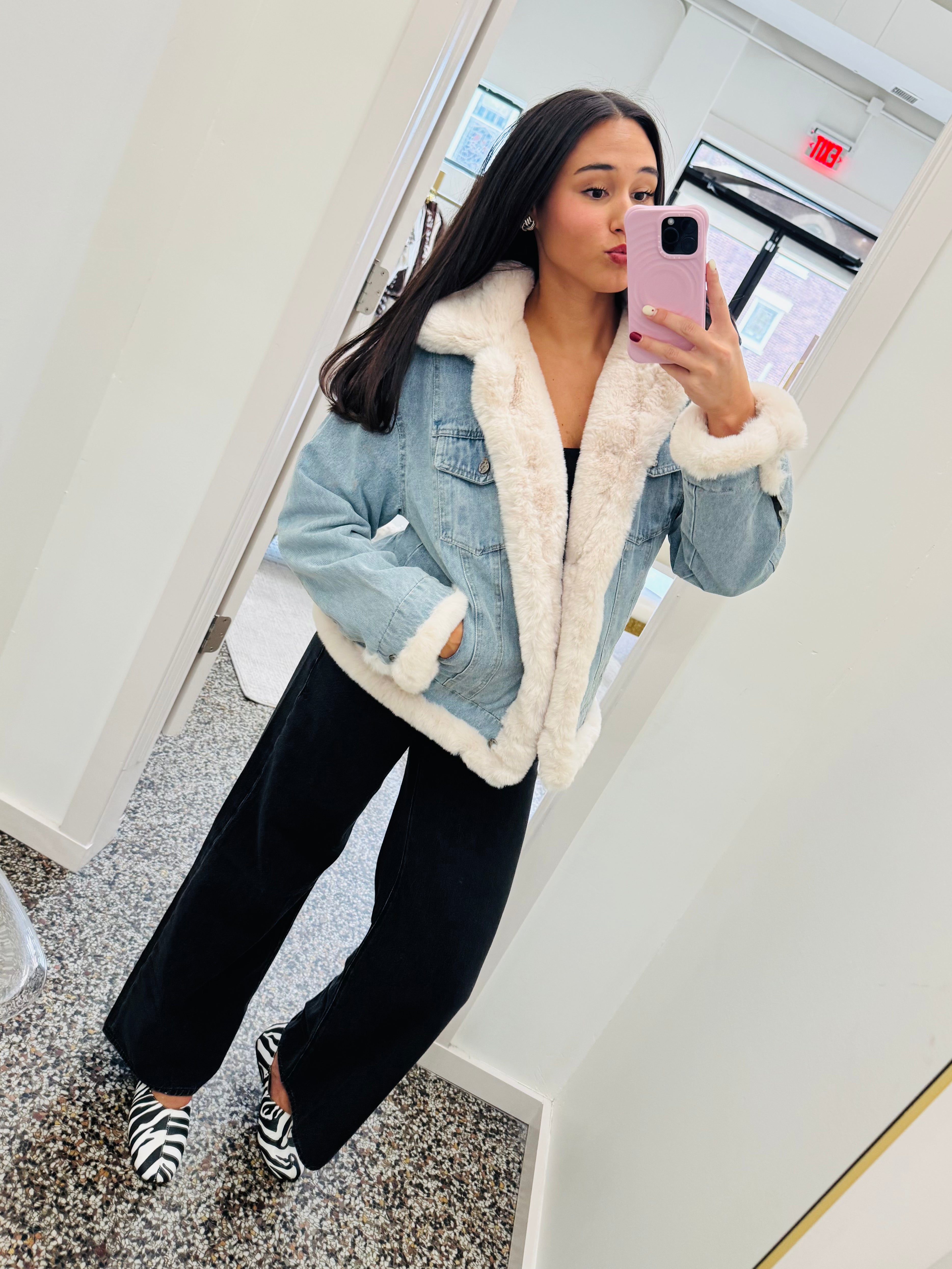 A woman takes a mirror selfie wearing a light blue denim jacket with a thick white sherpa lining, black wide-leg pants, and black-and-white striped shoes in a bright indoor space.
