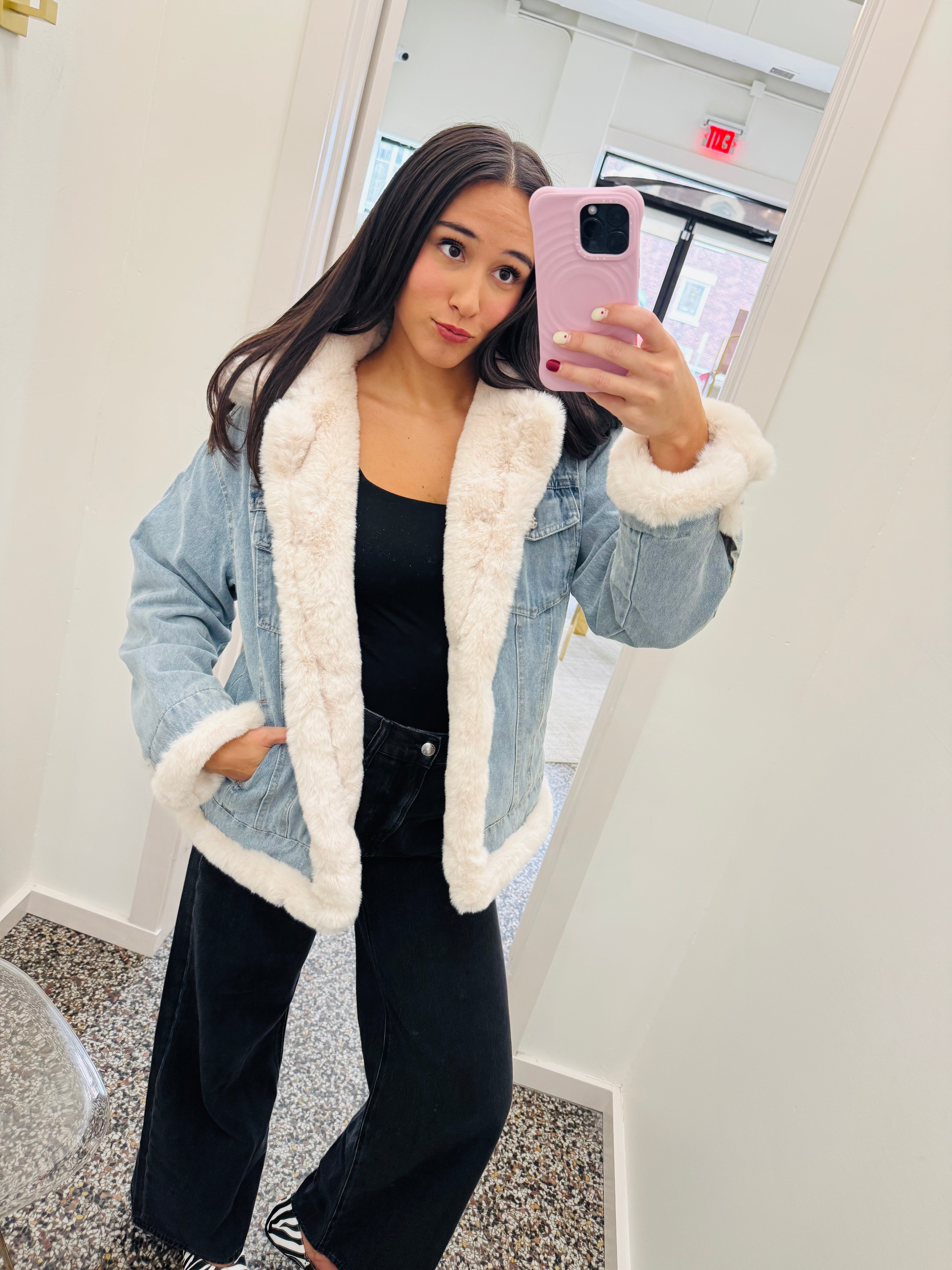 A woman takes a mirror selfie wearing a light blue denim jacket with a thick white sherpa lining, styled with a black tank top and black wide-leg jeans.