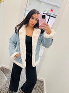 A woman takes a mirror selfie wearing a light blue denim jacket with a thick white sherpa lining, styled with a black tank top and black wide-leg jeans.