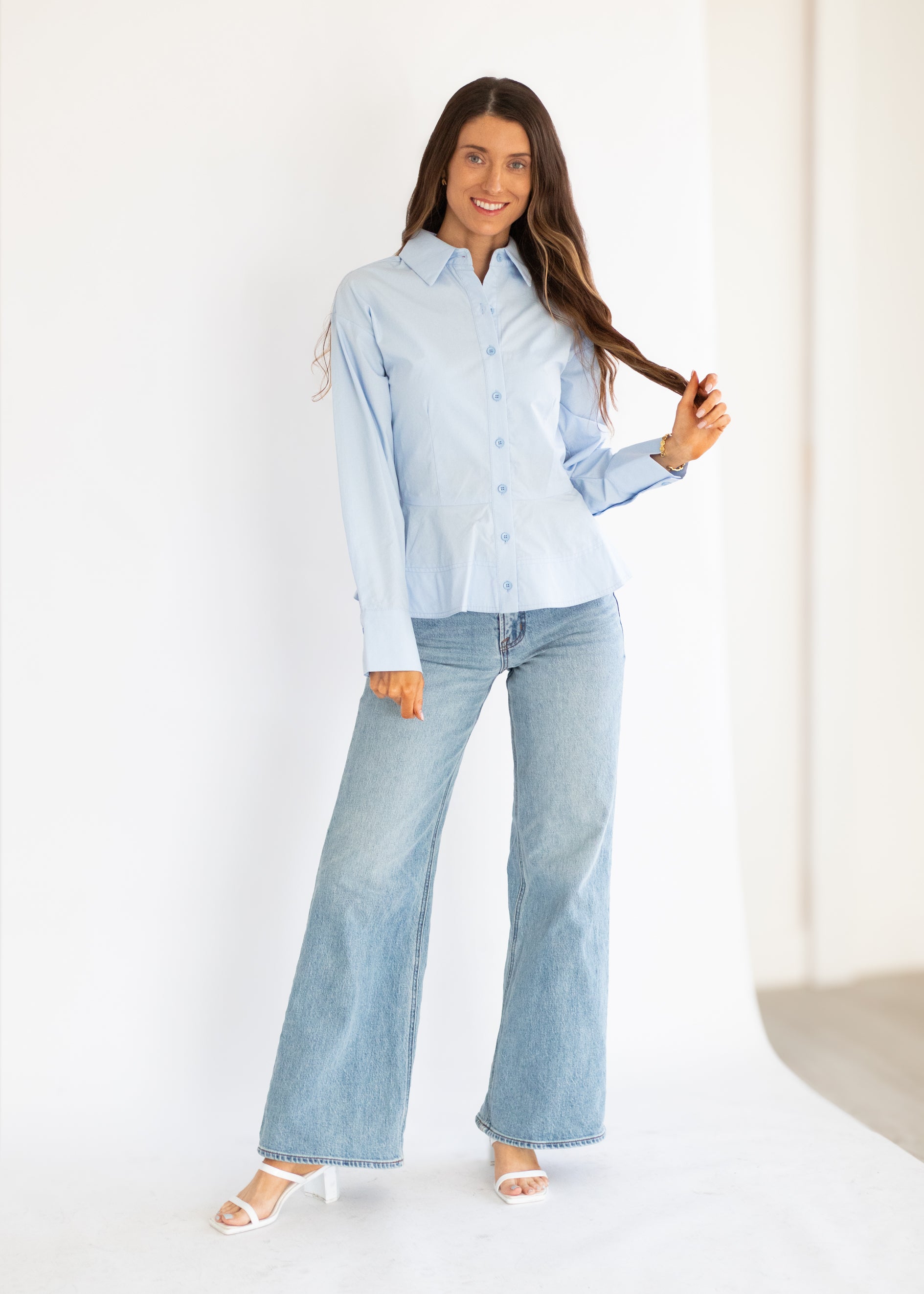 A woman modeling a light blue long-sleeve button-up shirt and matching light blue wide-leg jeans, standing against a clean white studio backdrop.