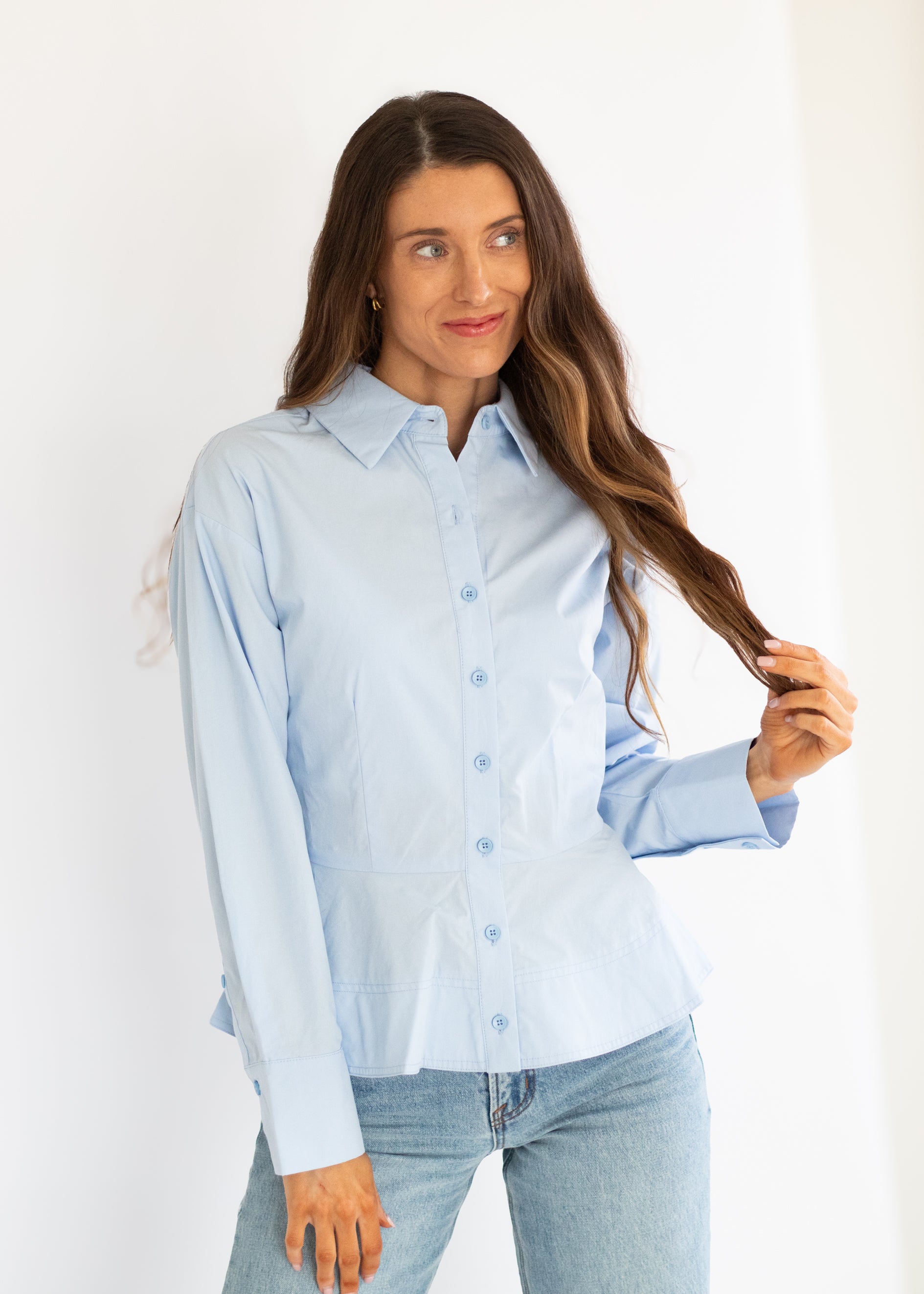 A woman modeling a light blue long-sleeve button-up shirt with light-wash blue jeans against a plain white studio background.